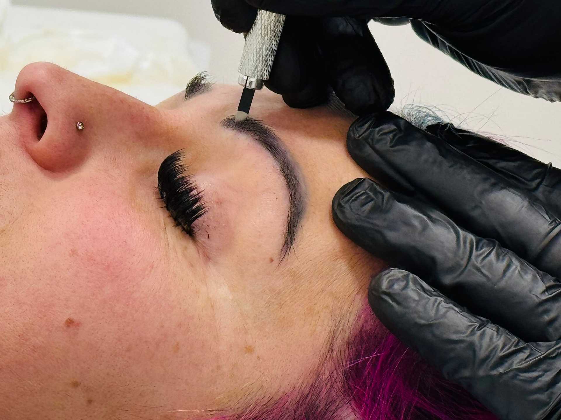 Person receiving eyebrow tattoo procedure with technician using a handheld tattoo tool.